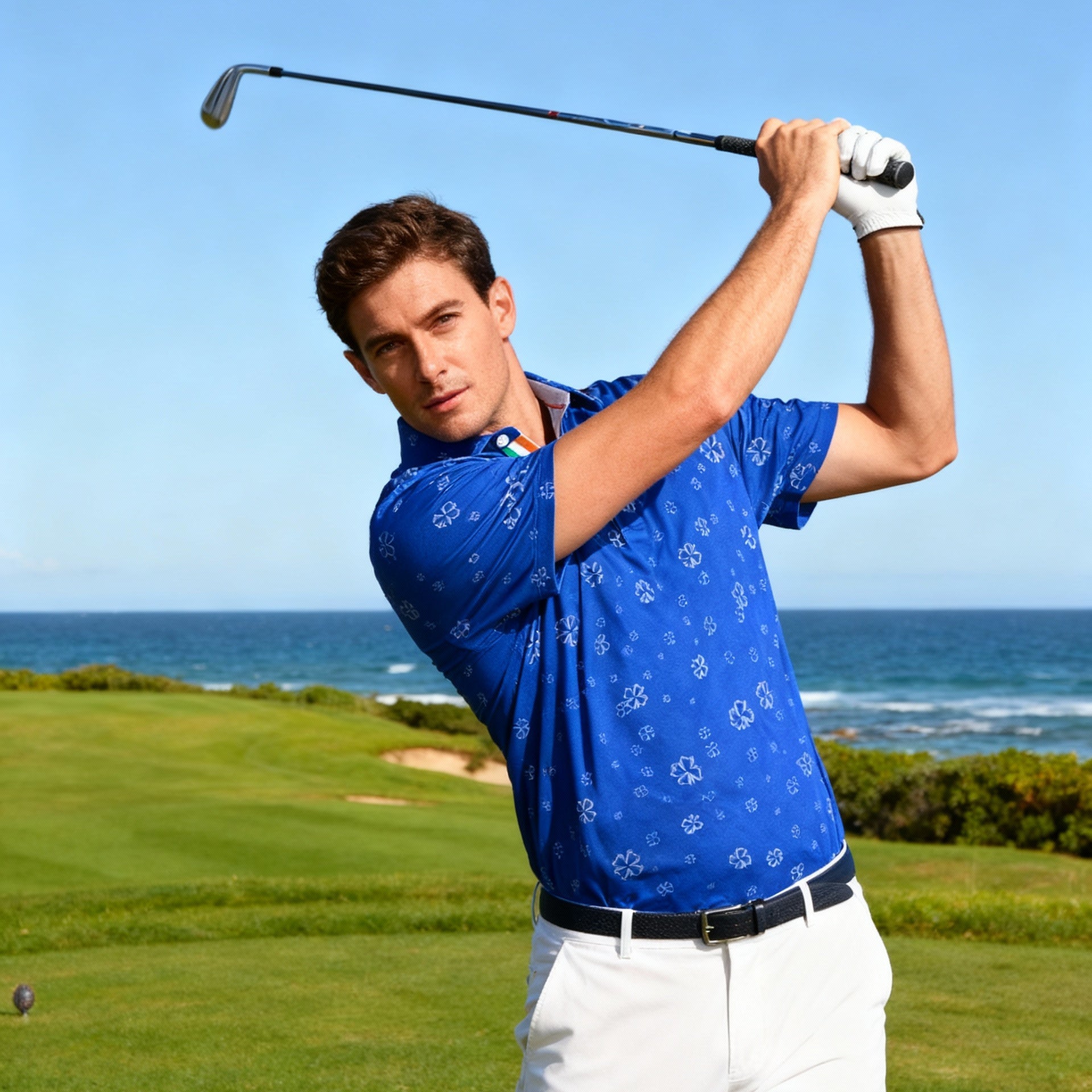 Man in blue shirt and white pants swinging a golf club on a golf course with ocean view.